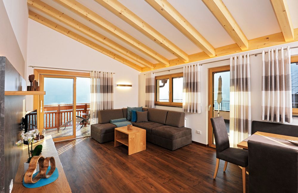 Modernes Wohnzimmer mit Holzbalkendecke, dunkelgrauer Couch und großen Fenstern mit Vorhängen