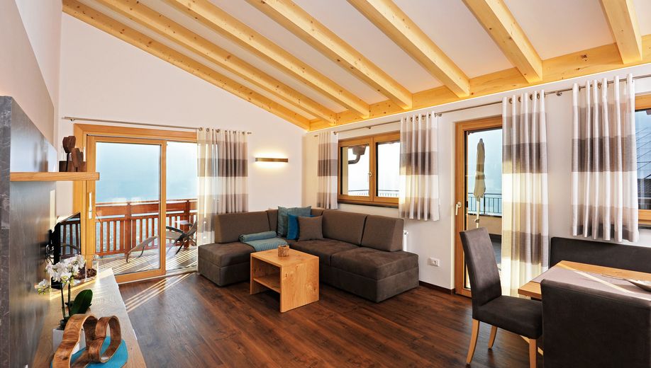 Modernes Wohnzimmer mit Holzbalkendecke, dunkelgrauer Couch und großen Fenstern mit Vorhängen