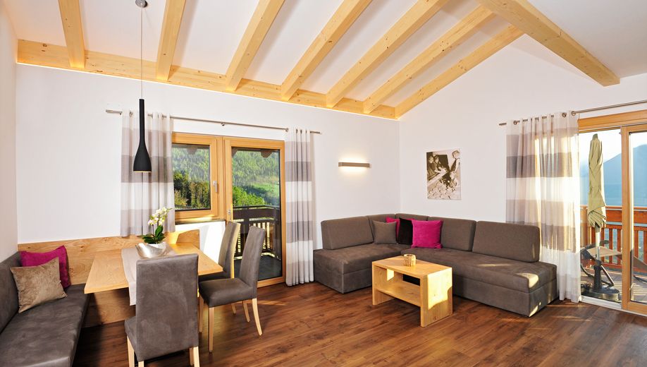 Helles Wohnzimmer mit Holzbalkendecke, Essbereich und Sofaecke mit bunten Kissen.