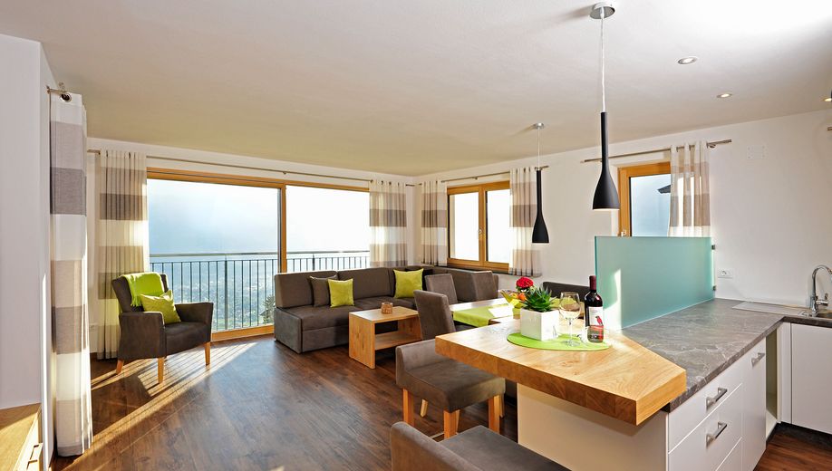 Helles Wohnzimmer mit Sofa, Sesseln, Holzboden und angrenzender Küche mit Bar und Fenster mit Blick ins Freie.