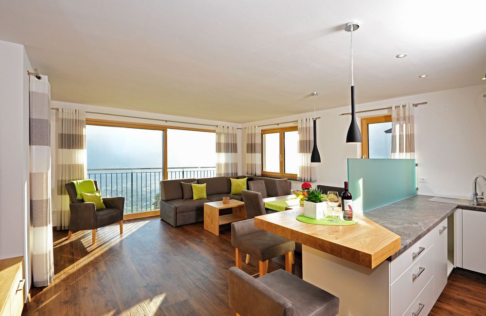 Helles Wohnzimmer mit Sofa, Sesseln, Holzboden und angrenzender Küche mit Bar und Fenster mit Blick ins Freie.