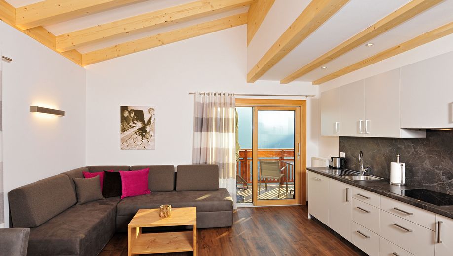 Modernes Wohnzimmer mit Holzbalkendecke, grauem Sofa, bunten Kissen und Küchenzeile mit Sicht auf Balkon