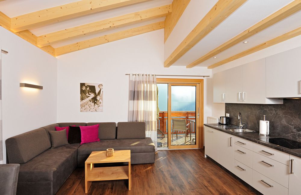 Modernes Wohnzimmer mit Holzbalkendecke, grauem Sofa, bunten Kissen und Küchenzeile mit Sicht auf Balkon